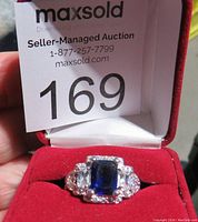 Front view of the ring showing the large blue rectangular gemstone center with smaller clear stones around it and red velvet background.