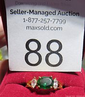Front view of size 8 gold tone ring with an oval green center stone and two clear marquise side stones, displayed on red velvet ring holder with auction lot tag.
