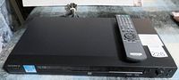 Sony DVD player with remote control placed on top. Front panel has power and playback buttons with disc tray.