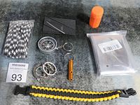 Survival gear items arranged on a dark surface showing rope, compass, whistle, compact folding wire saw, sealed space blanket, waterproof matches container, and paracord bracelet.
