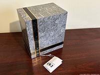 Full view of the cube-shaped urn showing grey fossil stone surface with black and gold stripe inlay and attached tag resting on wooden table.