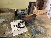 Wide shot of the Delta Duo Sander Grinder combo unit with sanding belts and manuals on wooden workbench.