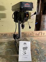 Front view of Jobmate 8 inch drill press on wooden workbench with instruction manual placed in front.