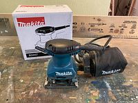 Makita palm sander with dust collection bag plugged in, showing front and side angle along with original box