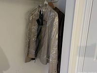 Khaki lightweight men's jacket with patches hanging in a closet covered in protective plastic with a large black umbrella behind it.