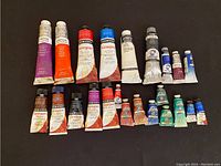 Top-down view of all oil paint tubes arranged on a dark surface showing various brands and colors.