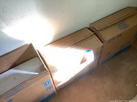 Three cardboard boxes containing new white Toto toilet tanks lined up on floor against wall.