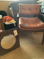 Chair with wooden frame and brown tufted seat and back cushions, alongside metal framed mirror on carpeted floor.