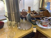 Wide view of large stacked aluminum pots, mugs, glass and crystal serving dishes, metal serving tray, wooden cutting boards, and a folding slicer on a table.