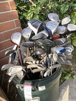 Overview of many golf clubs in a green trash barrel, heads and shafts visible with various brands including X-Cat and Carbite.