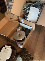 Box of metal cookware including baking sheets, mixing bowls, metal pots with lids and cast iron muffin pan