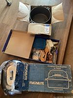 Photo showing large black cooking pot in a cardboard box, a box of assorted kitchen utensils, and a Magnalite branded covered oval roaster box with its shiny metallic lid resting on top.