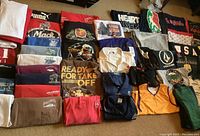 Top view of folded men's printed T-shirts and casual wear laid flat on carpet, showing variation of brands, colors, and graphics including Puma, Mack, and humorous prints.