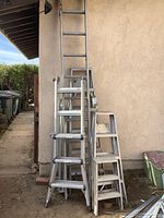Photo showing a group of metal ladders stacked against a wall outdoors including a tall extension ladder and shorter step ladders.