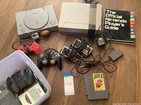 Wider lot overview showing Sony PlayStation 1 and NES consoles, controllers, game cartridge, official game guide, and additional cables and adapters.