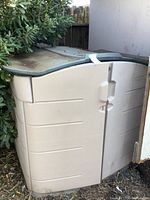 View of the beige plastic shed with gray lid partially surrounded by greenery and a white wall.
