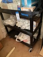 Front angled view of black plastic three-tier utility cart showing stacked shelves and wheels, with some items on the shelves for scale only, not included in sale.