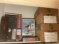 Shelf with boxed barbecue skewers, binocular microscope set, Scruples game, and two cardboard boxes labeled 'Poker Stuff' containing poker game accessories and board games.
