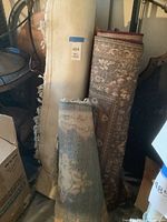 Three rolled area rugs leaning against a bed frame among other items in storage. One beige rug is frayed, one muted floral patterned rug, and one red ornate patterned rug.