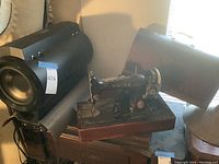 View showing black antique Singer sewing machine with gold name and decorative detailing on wooden base, alongside black cylindrical modern subwoofer speaker with control knobs.