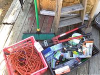 Photo of lot items showing bolt cutters, electric cord coiled in pink crate with various small packaged items, broom with green handle and black bristles, wooden step ladder, and shovel with wooden handle.