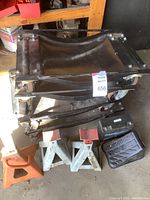 View showing four black metal wheel dollies stacked, two orange jack stands, one pair gray stands, a small black battery, and wheel braces.