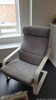 Full view of adult IKEA Poäng chair by window showing gray fabric cover and white wooden frame with curved armrests.