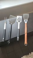 Four BBQ tools laid out on counter, showing black and wooden handles, metal heads including forks and spatulas with slotted holes