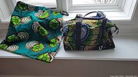 The purse and matching drawstring fabric bag displayed on a window sill, showing the bold multicolor circular African print, leather accents, and details.