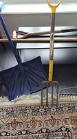 Photo showing blue large snow shovel with wooden handle and earth turning fork with yellow marked handle leaning against a table.