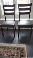 Two wood dining chairs with ladder-back design and cushioned faux leather seats placed side by side.