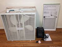 Photo showing box fan, portable heater with instruction manual, and sealed air filter package against wall on wood floor