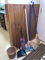 Overview photo of cleaning supplies including broom, mop, dustpan, feather duster and small basket arranged against wood panel wall