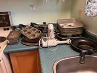 Photo of multiple cookware items including pots with lids, muffin tin, loaf pans, frying pans, a hand mixer, and electric griddle on kitchen counter.