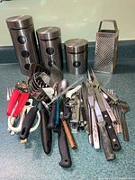Photo shows an assortment of kitchen cutlery including knives, spoons, forks, a cheese grater, and three stainless steel containers in varying sizes.