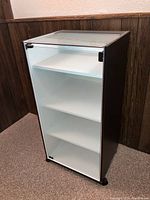 Front and angled view showing white interior, four shelves, glass front door with magnetic latch, and caster wheels on bottom.