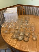 Photo showing all glassware items arranged on a round wooden table including wine glasses, beer mugs, tumblers with branding, mugs, and assorted smaller glasses with a white storage box for the wine glasses.