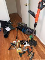 Full view showing rechargeable weed wacker, electric blower, garden sprinkler, extension cords, and assorted tools spread on floor.