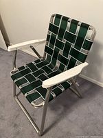 Front angled view of one green woven strap lawn chair with white armrests and silver tubular metal frame against plain wall