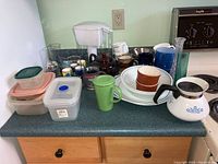 Wide view of assorted kitchen ware on a counter including CorningWare pieces, mugs, glasses, tea pots, and storage containers.