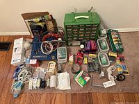 Wide shot showing a large assortment of nails, fasteners, cords, tapes, switches and related DIY supplies arranged on carpet and hardwood floor. Includes green metal drawer organizer, multiple rolls of tape, various electrical cords, screws, wires, sandpaper, electrical outlets and covers.