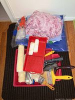 Top view of assorted painting supplies including paint brushes, rollers, red paint trays, and fabric drop sheets stacked together