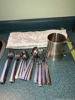 Items neatly arranged on counter: cutlery including forks, knives, spoons, white folded table cloth behind them, pewter mug next to cutlery.