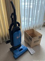 Full view of blue Hoover Tempo upright vacuum cleaner standing near window with beige curtains and cardboard box beside it.