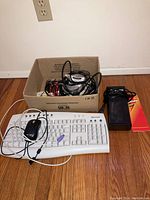Photo of white Microsoft wired keyboard with PS/2 connector, black wired mouse, and boxed battery charger on floor beside a wall outlet