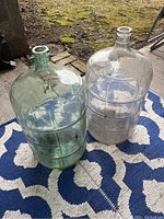 Two large glass jugs placed outdoors, one clear and one greenish tint, showing surface dirt.