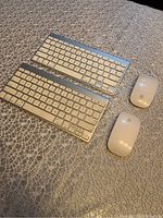 Two Apple wireless Magic Keyboards and two Apple Magic Mice placed on a textured fabric surface.