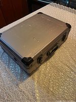 Closed view of dark grey metal briefcase with ribbed surface and reinforced black corners showing front metal latches and handle.