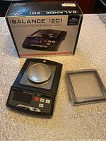 Digital precision balance scale with stainless steel tray, buttons and display visible beside transparent protective cover and original box on table.