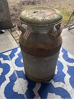 Front view of large metal milk jug showing cylindrical body, side handles, lid, and wear.